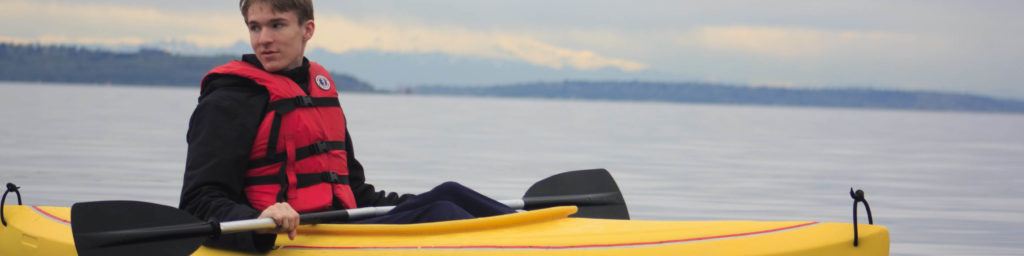 Young person in a red and black life jacket sitting in a yellow kayak on a calm lake or bay with forested mountains in the background, promoting church summer camps and water activities.