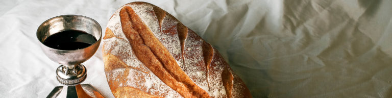 Religious communion elements: an ornate silver chalice filled with dark red wine and a freshly baked loaf of bread dusted with flour on white linen cloth