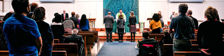 Church worship service photo with congregation members standing in the sanctuary and choir members in colorful robes on stage