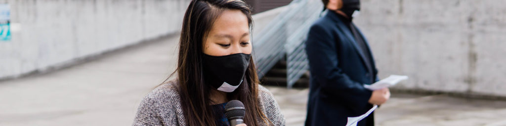 Woman wearing black mask speaking into microphone at an outdoor event.