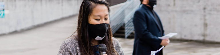 Woman wearing black mask speaking into microphone at an outdoor event.