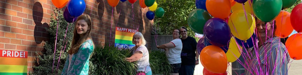 HAPPY PRIDE and PRIDE! text with First Church branding, people standing with colorful balloons and pride flag decorations at church event.