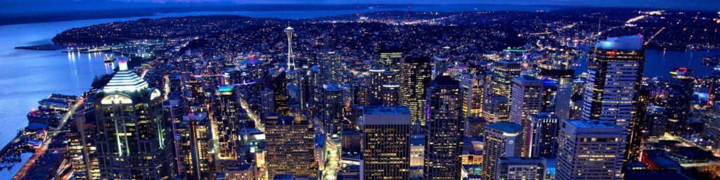 Seattle cityscape at dusk showing illuminated downtown buildings, Puget Sound with reflections, and the surrounding urban landscape.