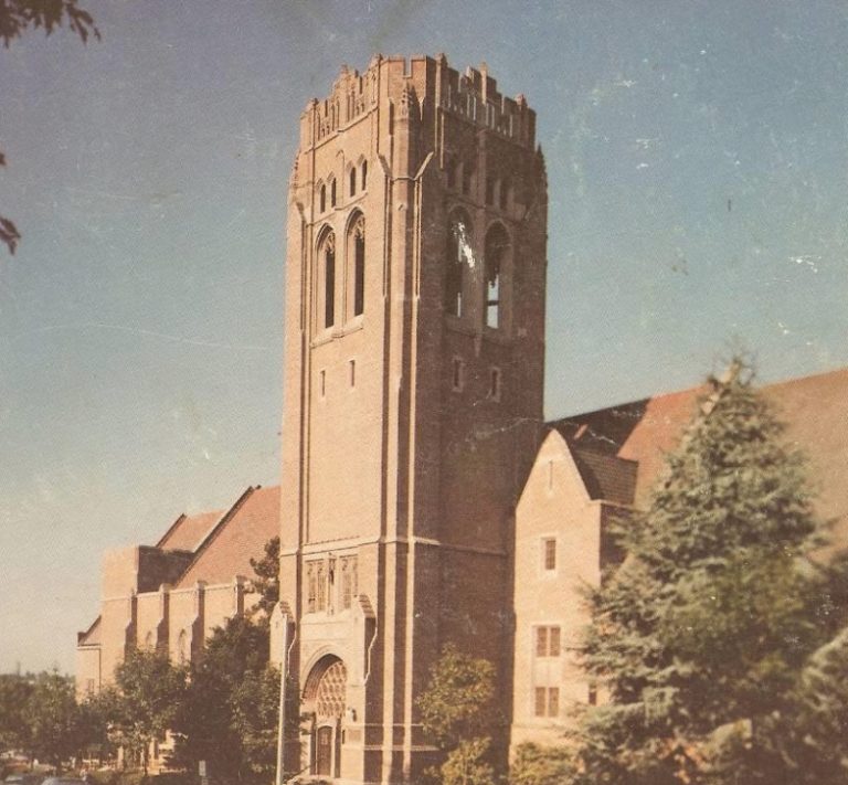 Historic church building with tall brick tower and art deco architectural style, surrounded by evergreen trees and residential buildings.