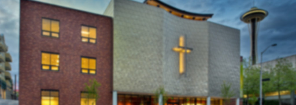Photo of First Church Seattle building exterior at twilight with red brick structure, illuminated windows, golden cross on white wall, and Space Needle visible in background.