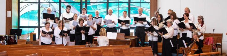 Church choir members in white and black formal attire singing with hymnals in a bright sanctuary with blue stained glass windows and wood paneling.