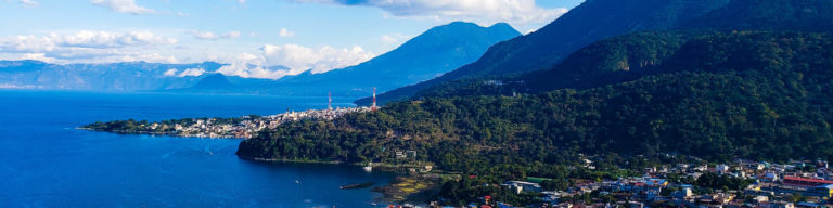Aerial photograph of a mountainous tropical coastline with turquoise water, lush green hillsides, and a coastal town nestled against dramatic peaks under blue sky with white clouds