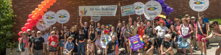 Large group photo of church community members at pride event holding signs reading 'All Are Welcome - Reconciling Congregation of The United Methodist Church' and 'pride + faith' signs, with colorful balloon arch and rainbow pride flags.