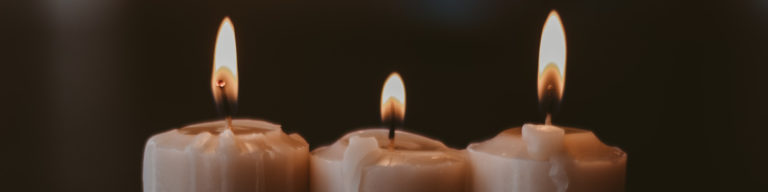 Three lit candles with flames against a dark background, representative of contemplation, prayer, or memorial.