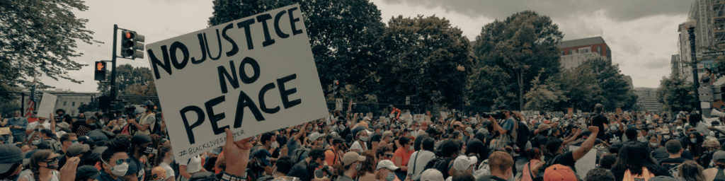 Photograph of a large crowd at a protest rally with a prominent sign reading 'NO JUSTICE NO PEACE' and '#BLACK LIVES MATTER' visible, representing a social justice demonstration.