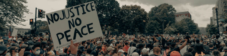 Photograph of a large crowd at a protest rally with a prominent sign reading 'NO JUSTICE NO PEACE' and '#BLACK LIVES MATTER' visible, representing a social justice demonstration.