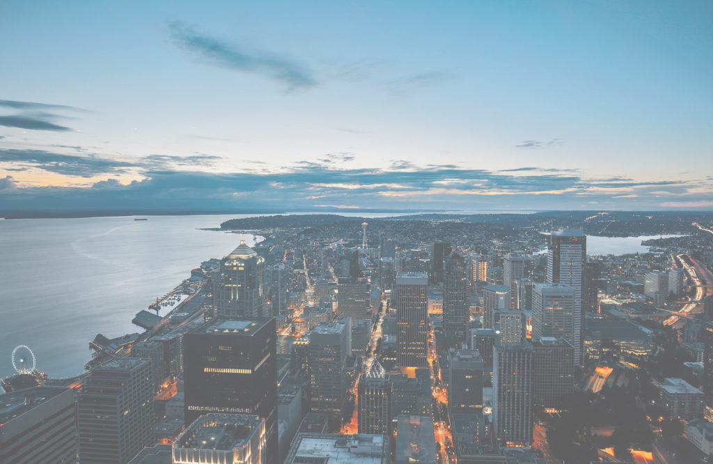 Aerial evening photograph of Seattle skyline at dusk showing illuminated downtown buildings, Puget Sound waterfront with boats, and surrounding residential neighborhoods with forested areas.