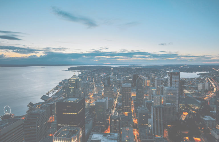 Aerial evening photograph of Seattle skyline at dusk showing illuminated downtown buildings, Puget Sound waterfront with boats, and surrounding residential neighborhoods with forested areas.