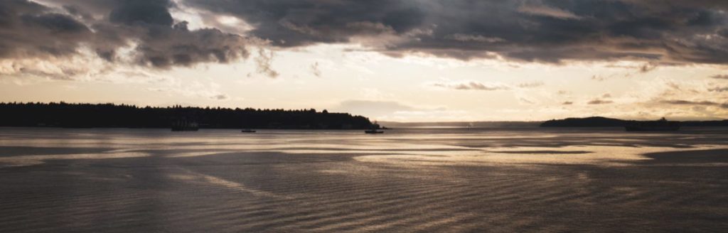 Serene waterfront landscape at dusk with calm water, forested shoreline, and dramatic cloudy sky reflected in the water.