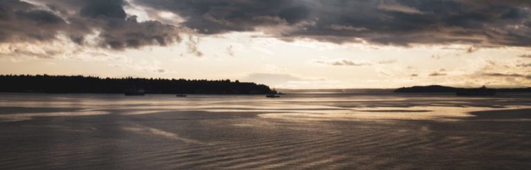 Serene waterfront landscape at dusk with calm water, forested shoreline, and dramatic cloudy sky reflected in the water.