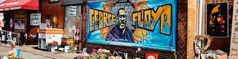 Street art mural featuring a portrait with the text 'PEACE FLOYD' in bold yellow and blue lettering, honoring George Floyd, with flowers and memorial items at the base.