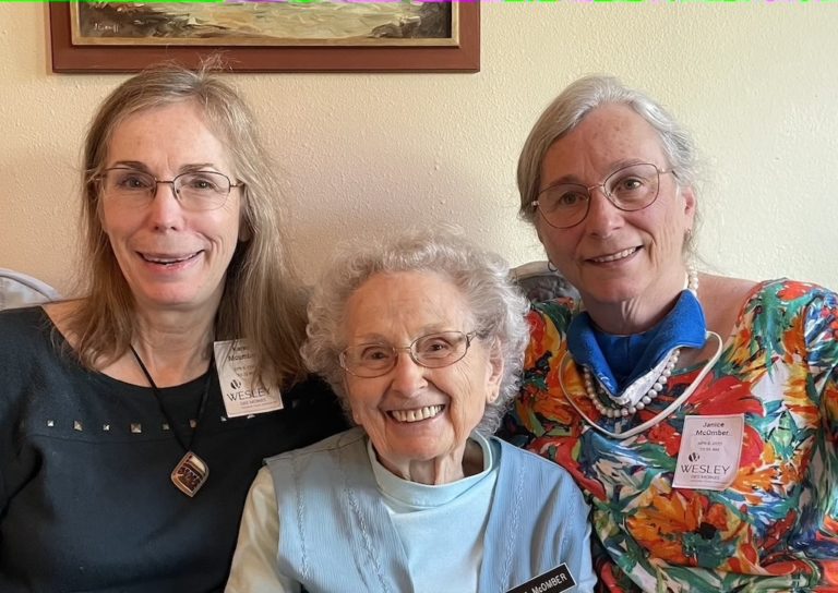 Three smiling women standing together, two wearing name tags, in an indoor setting.