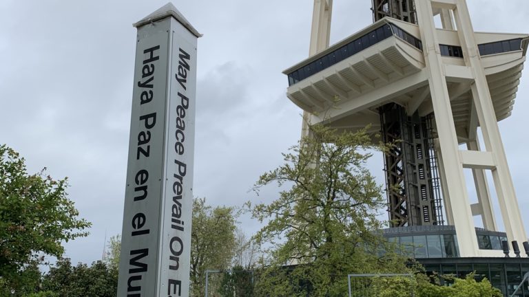 Peace Pole monument with vertical white pillar displaying text "Haya Paz en el Mundo" and "May Peace Prevail On Earth" in multiple languages, next to modern white architectural building.