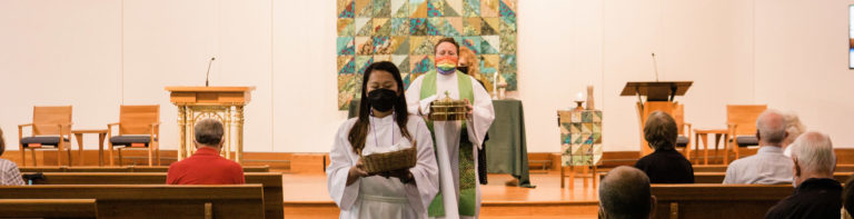 Church sanctuary service with congregants seated in wooden pews and two women in masks at the altar, one holding a Bible and the other in green vestments.