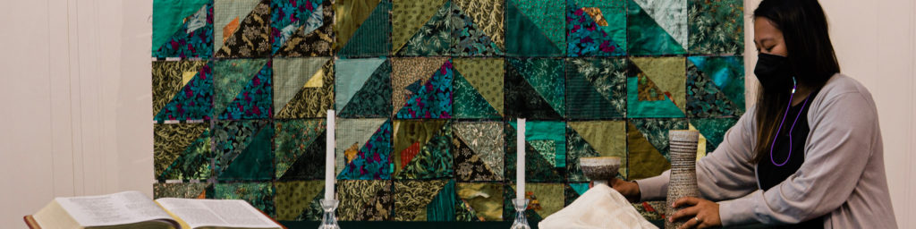Woman in light gray sweater with purple stethoscope around neck standing next to colorful patchwork quilt wall hanging with fabric samples.