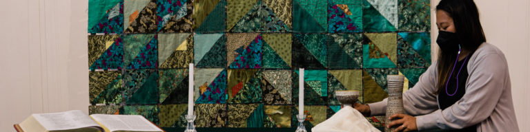 Woman in light gray sweater with purple stethoscope around neck standing next to colorful patchwork quilt wall hanging with fabric samples.
