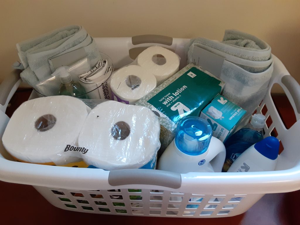 Close-up of bathroom essentials and hygiene products including rolled toilet paper, hand lotion, and other supplies in white and clear storage containers.