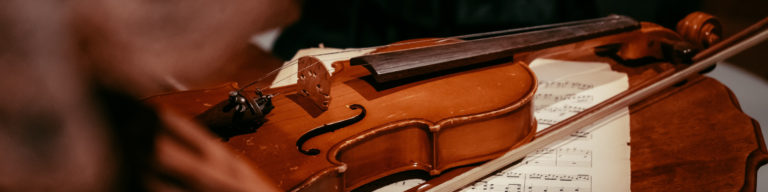 Close-up photograph of a wooden violin with sheet music, featuring a polished reddish-brown instrument and bow.