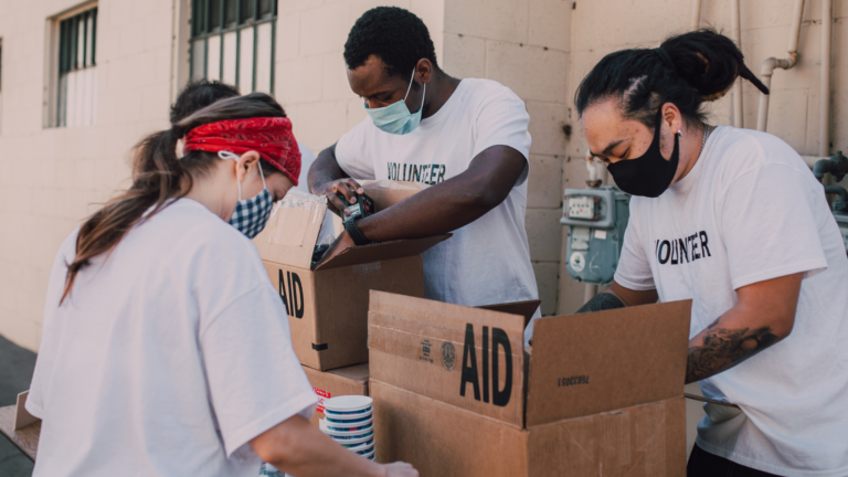 Group of volunteers wearing white shirts labeled 'VOLUNTEERED' and 'WINNER' assembling aid boxes together during a community service event.