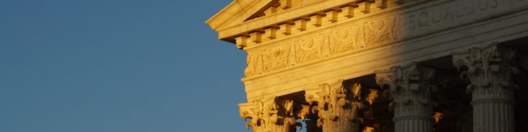 Close-up of the ornate columned facade of the United States Supreme Court building with golden classical architecture details against a blue sky