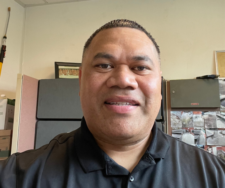 Portrait headshot of a man smiling at the camera in an indoor office or home setting with wall and framed pictures visible in background.