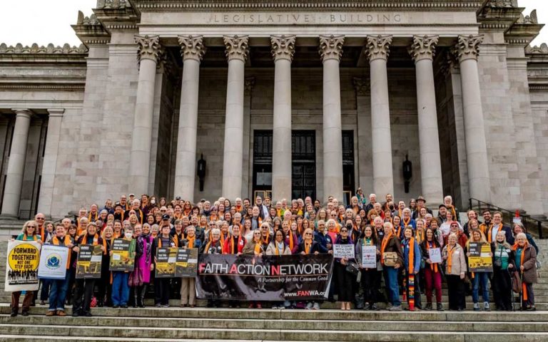 Large group photograph at the Legislative Building showing Faith Action Network (FAN) IFAD 2023 members, most wearing orange and gold garments, holding signs and a banner reading 'FAITH ACTION NETWORK A Partnership for the Common Good' with 'www.FANWA.org' visible.