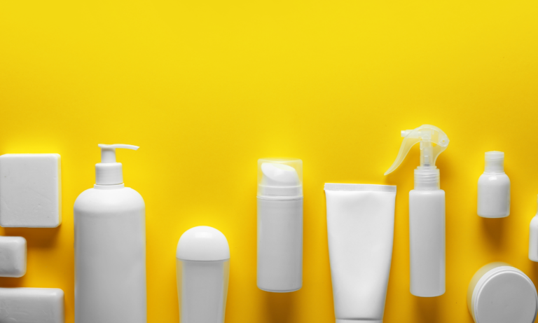 Minimalist still life photograph of white cosmetic and personal care product bottles and containers arranged on a bright yellow background, including pump bottles, spray bottles, and jars.