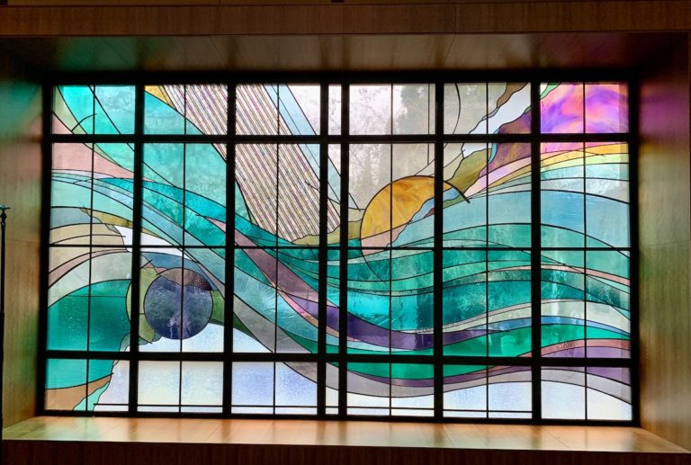 Modern stained glass window with geometric abstract design featuring turquoise, purple, yellow, and white glass panels arranged in a grid pattern with flowing curved lines and sun imagery representing contemporary church sanctuary art.