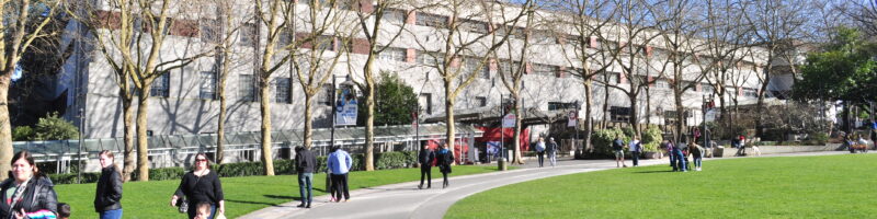 People strolling along a path through the green lawns of Seattle Center on a sunny day, with the Armory building and the Space Needle in the background