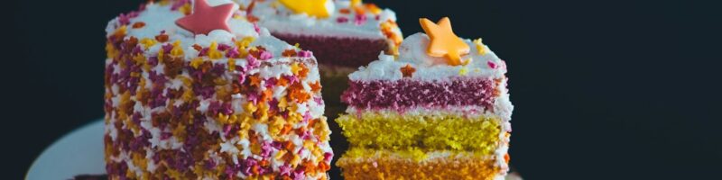 A sliced rainbow layer cake with white frosting and pastel fondant stars, displayed on a floral cake stand against a dark background