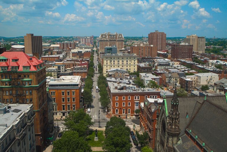Aerial view of dense rowhouse blocks in Baltimore under bright sun, with few trees between the rooftops.