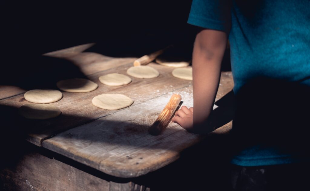 A child prepares dough on a rustic wooden table in a sunlit kitchen.