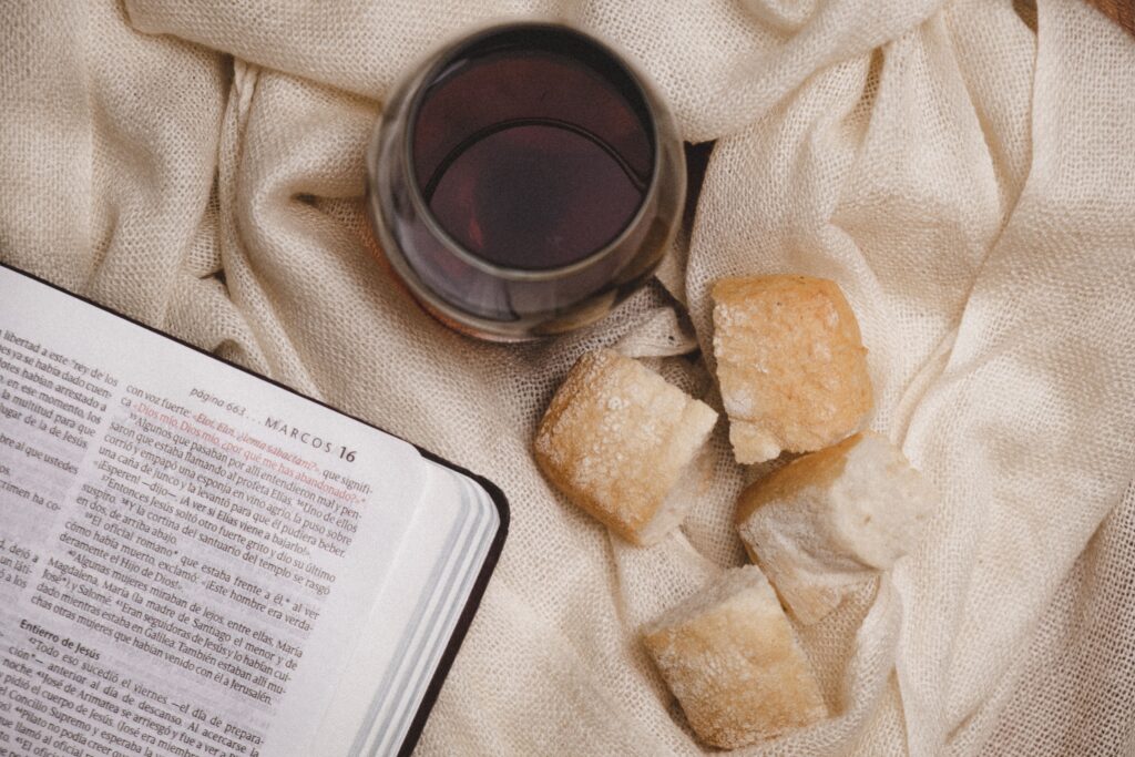 Overhead shot of a Bible, a glass of wine, and bread on a textured cloth.